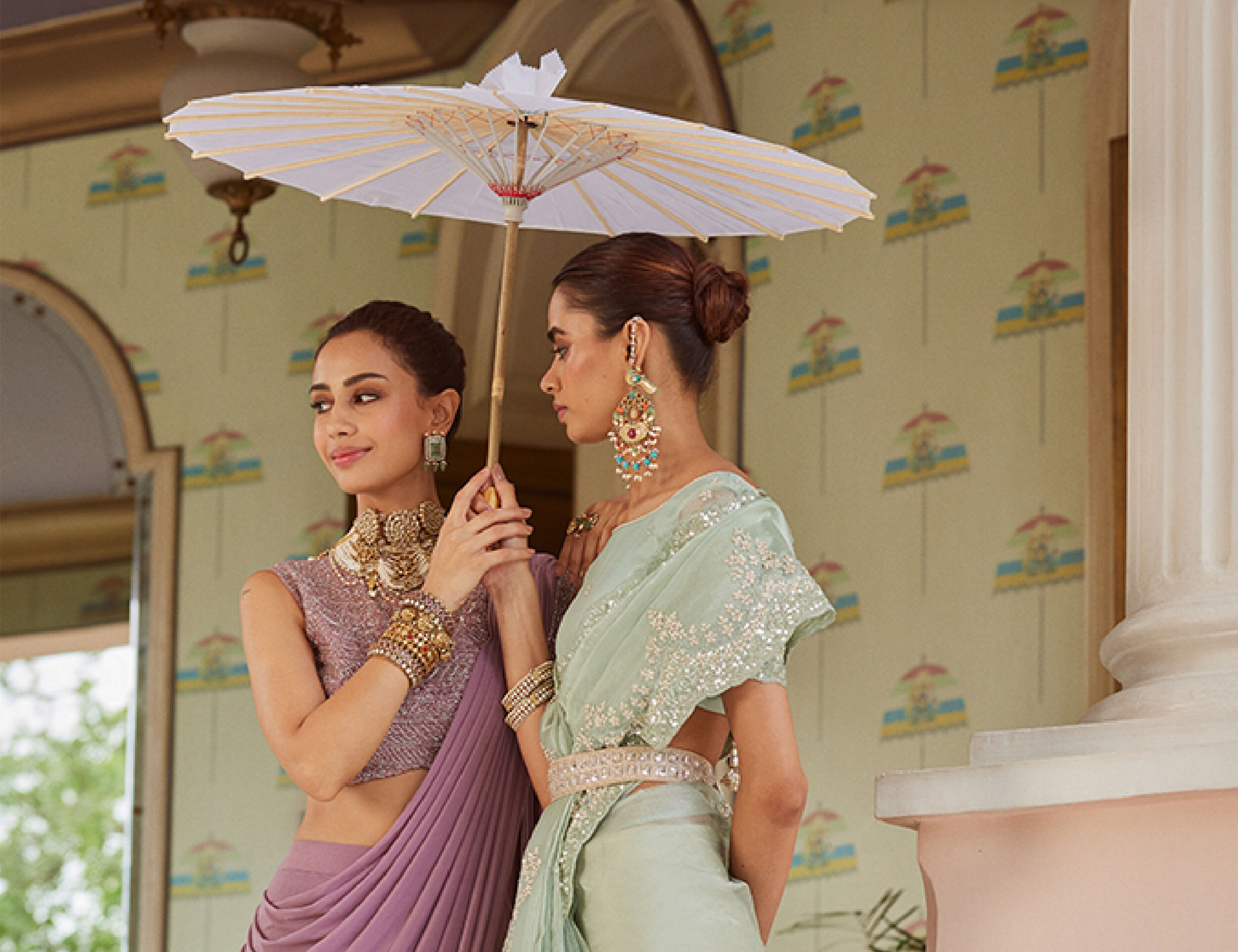 Two women wearing elegant designer sarees in pastel shades, accessorized with heavy traditional jewelry, standing under a white umbrella at a heritage-style venue.